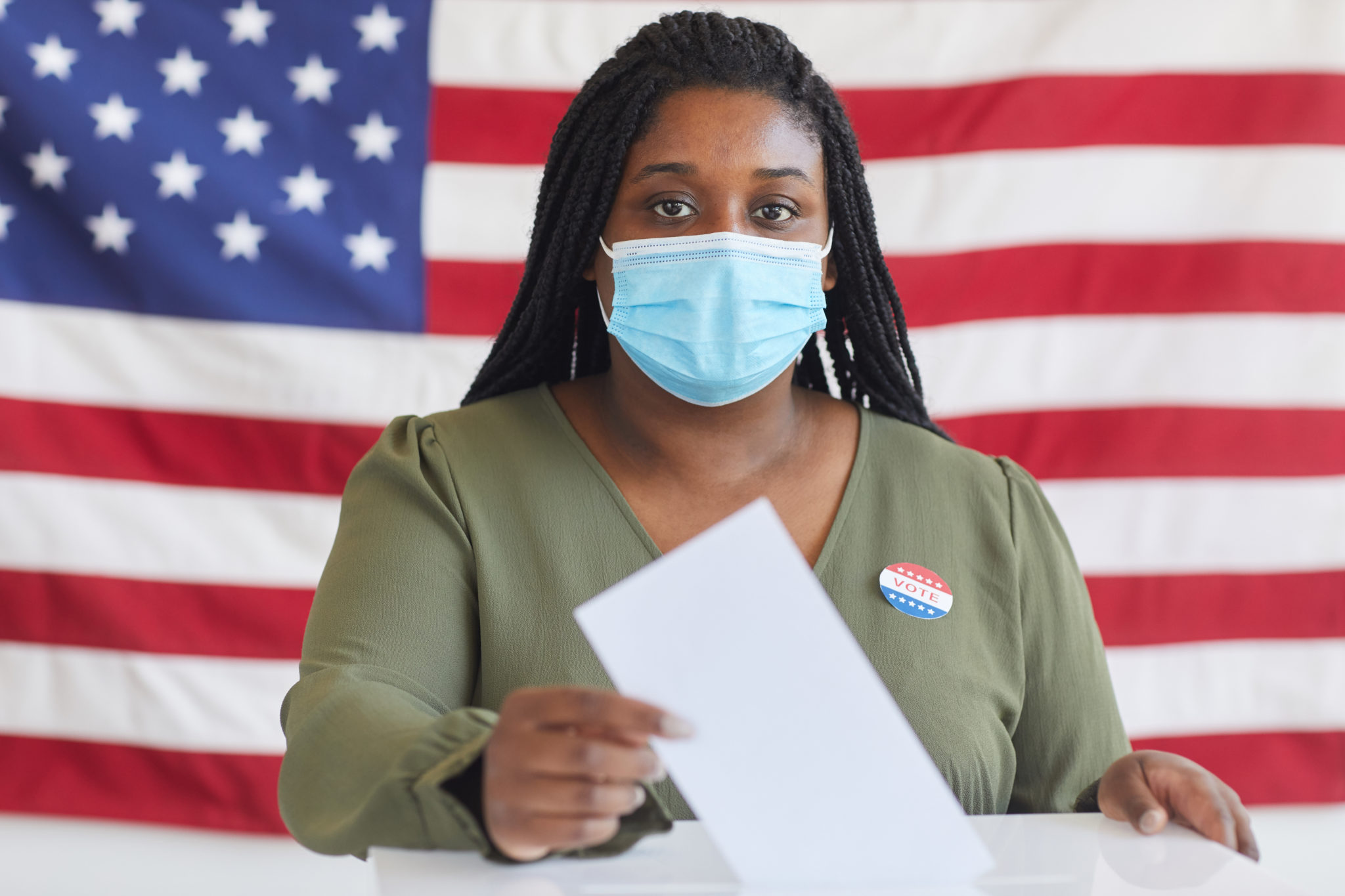 Portrait of young African-American woman wearing mask putting vote bulletin in ballot box and looking at camera while standing against American flag